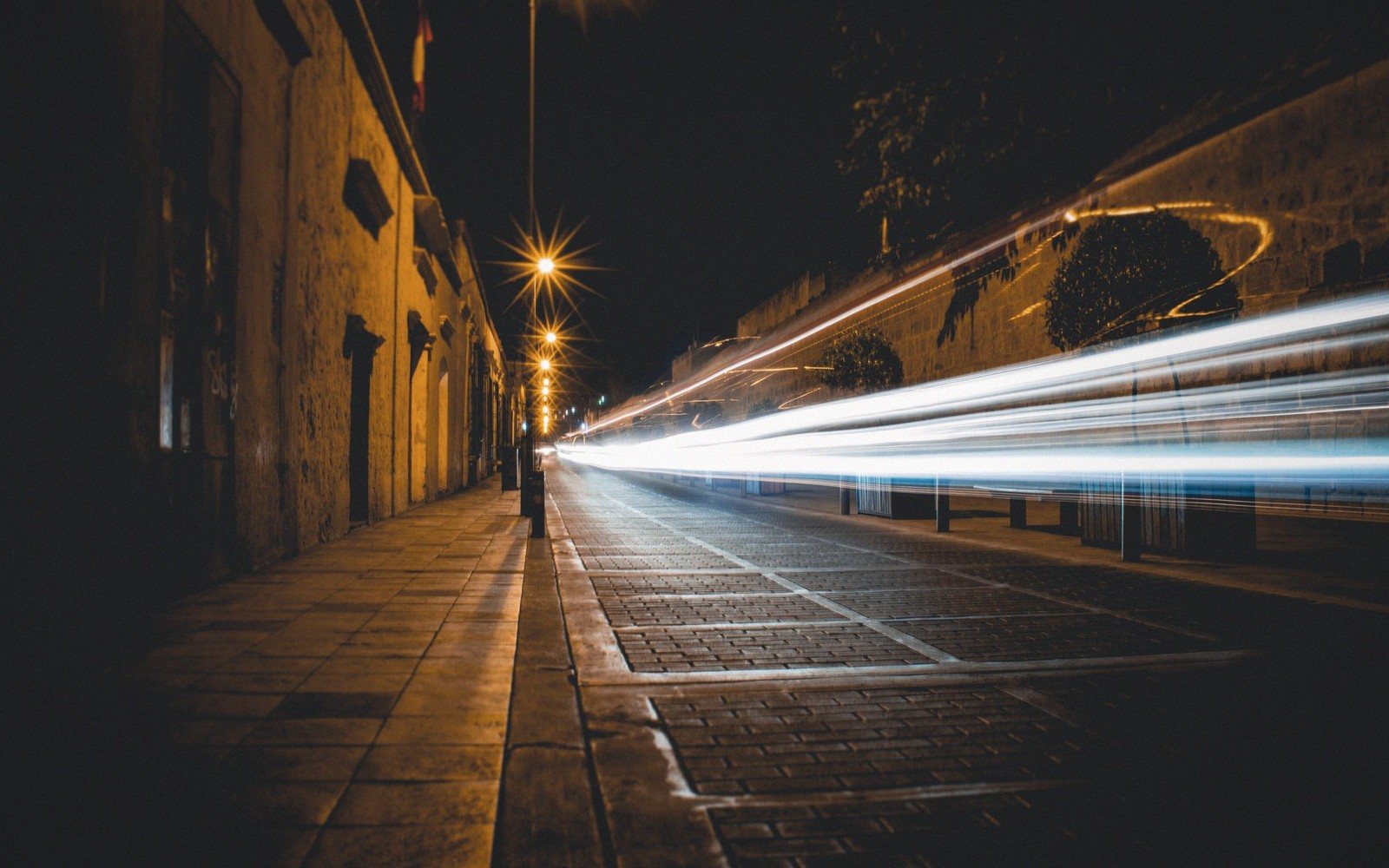 Long-exposure light trails streak down a cobblestone street beside historic stone buildings at night